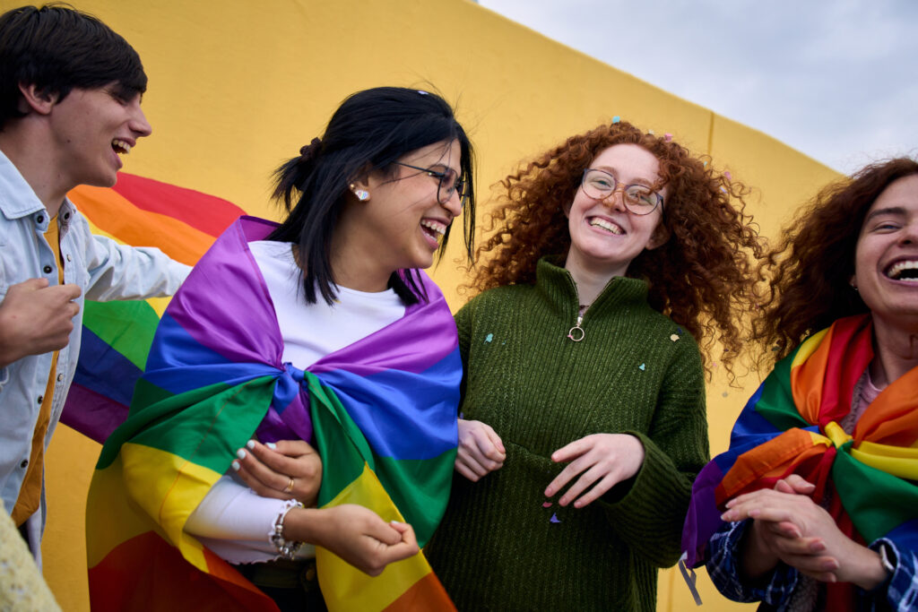 Young people of different backgrounds laughing in front of a yellow wall, with rainbow flags
